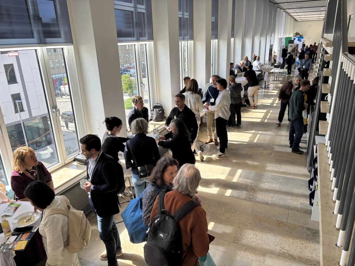 Teilnehmende einer Fachkonferenz tauschen sich in einem lichtdurchfluteten Foyer bei Kaffee und Gesprächen aus.