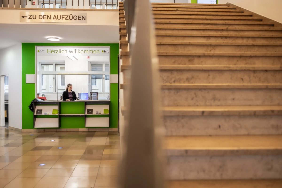 Empfangsbereich mit Mitarbeiterin hinter grün-weißer Infotheke im Gebäude des HDT.